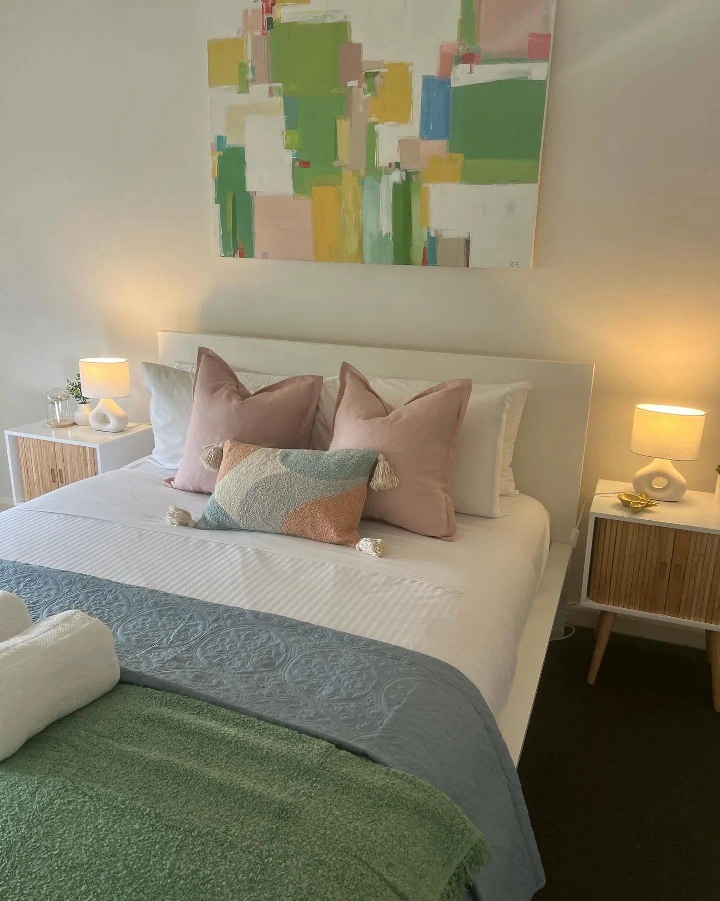 A neatly made bed with white sheets, pink and beige decorative pillows, and a multicolored throw blanket. There are two bedside tables with lamps on each side of the bed. A colorful abstract painting hangs on the wall above the bed.
