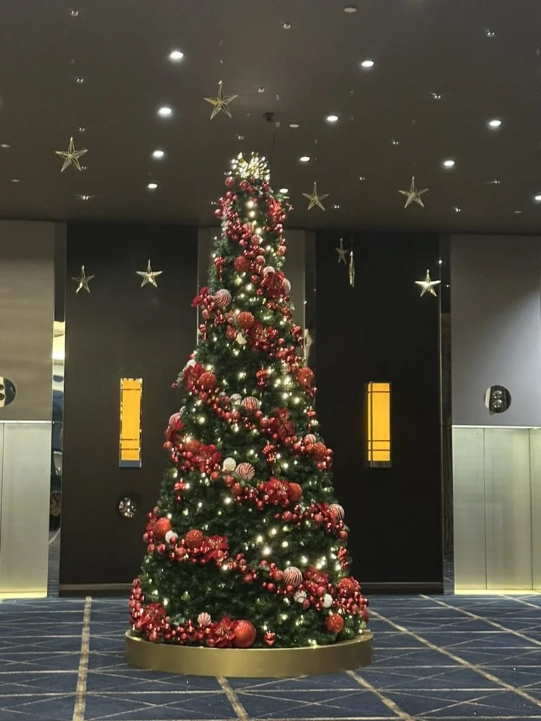 Christmas tree decorated with red and silver ornaments, garlands, and lights, at the Sofitel Melbourne. Gold star decorations hanging from the ceiling, and an elevator in the background.