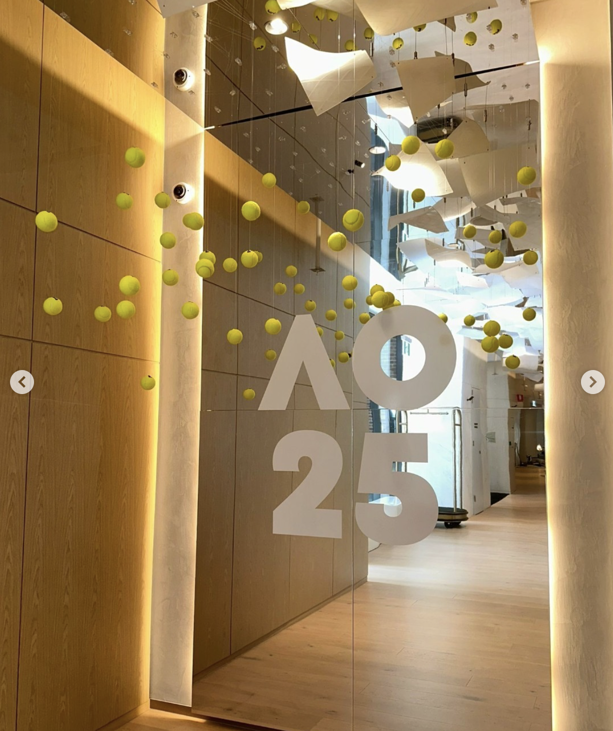 Interior of a modern building with a wooden wall and hanging yellow tennis balls, white paper sculptures, and the number '025' in large white font on a glass surface.