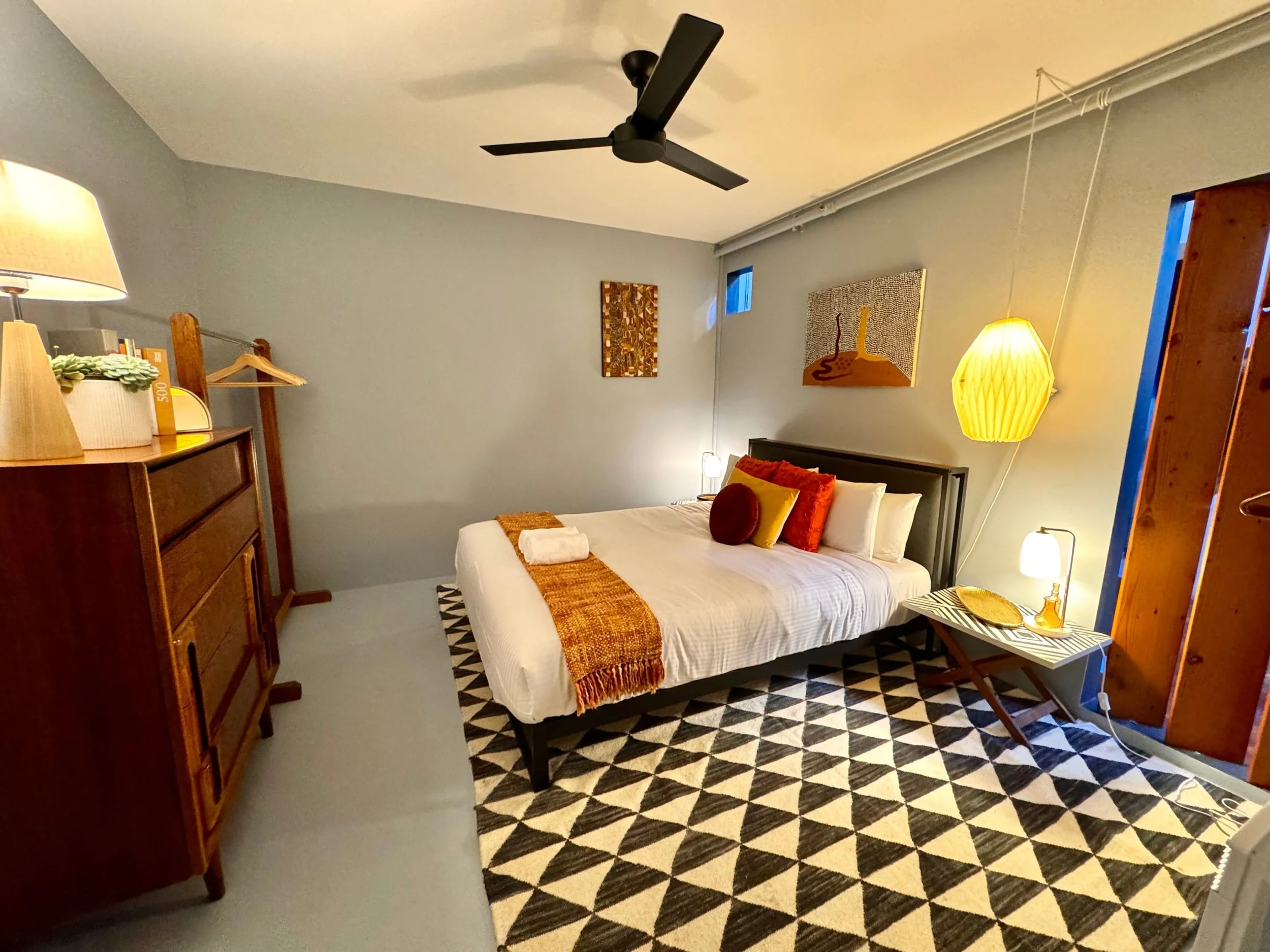 A cozy bedroom with a black bed frame, white bedding, and orange and red pillows. There are two paintings on the light gray wall, a wooden dresser with a lamp and plant, and a yellow pendant lamp hanging from the ceiling. A patterned black and white 