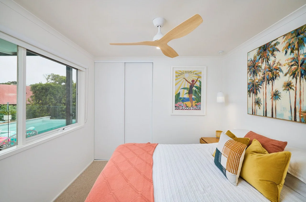 A bedroom with a window showing a pool outside, decorated with colorful pillows, tropical artwork, and white walls.