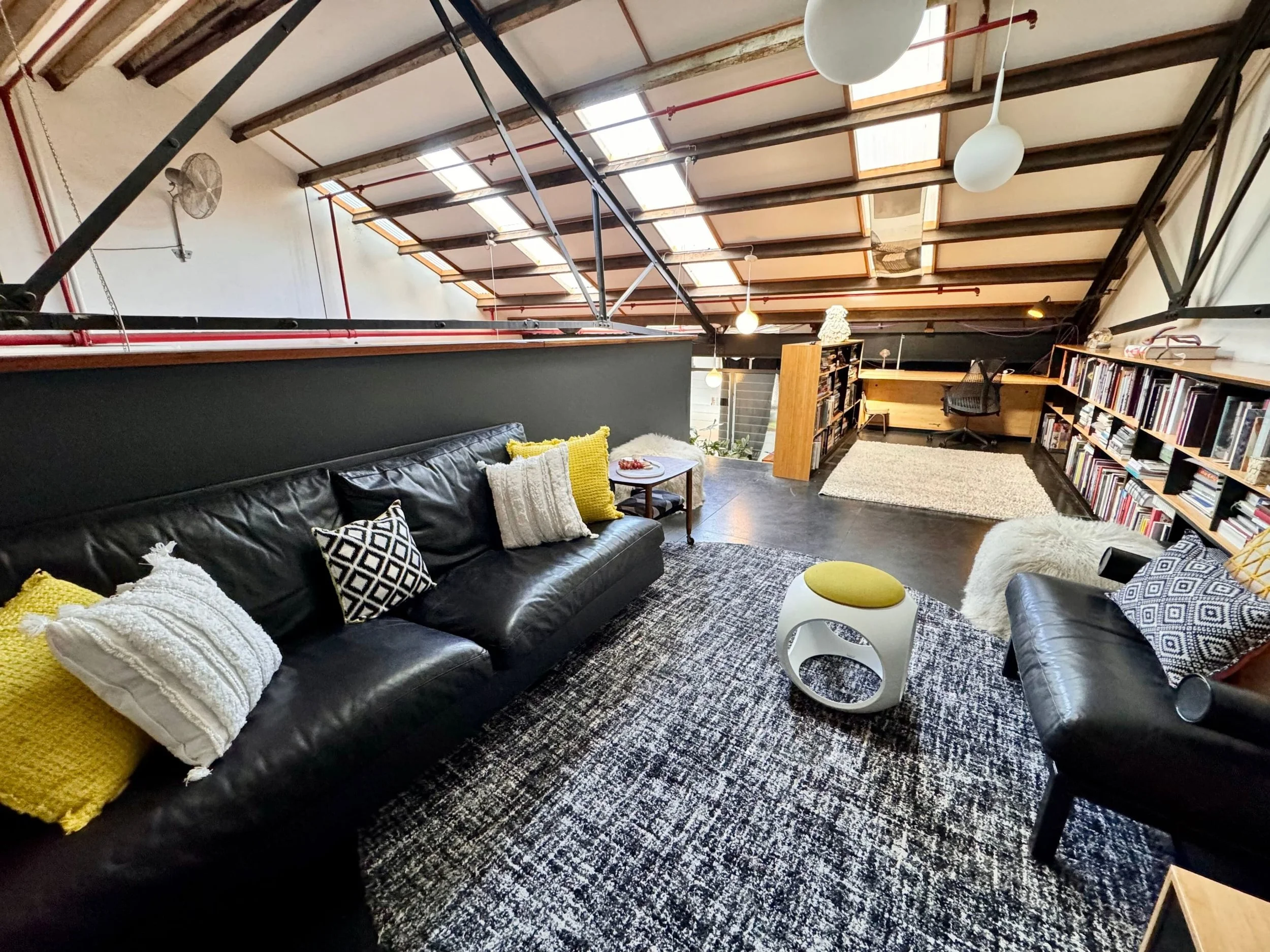 A stylish living room with a black leather sofa, decorative pillows, and a white side table, featuring a high ceiling with skylights, wooden beams, and eclectic décor including bookshelves, ceiling fans, and modern pendant lights.