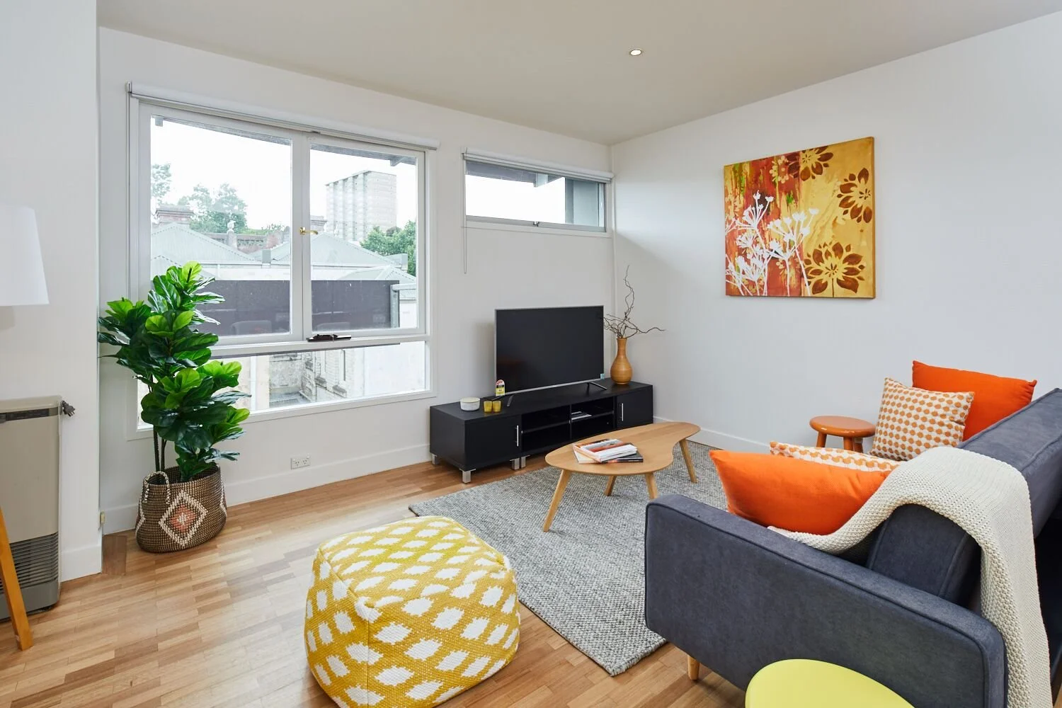 Bright living room with large windows, a gray sofa with orange and patterned pillows, a circular yellow and white pouf, a wooden coffee table with books, a TV on a black stand, a beige vase with twigs, a colorful floral painting on the wall, a green 