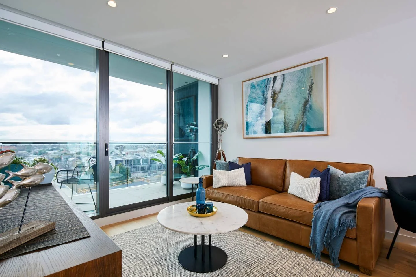 Modern living room with a leather sofa, throw pillows, a white marble coffee table with blue vases, and large balcony windows showing a city view.