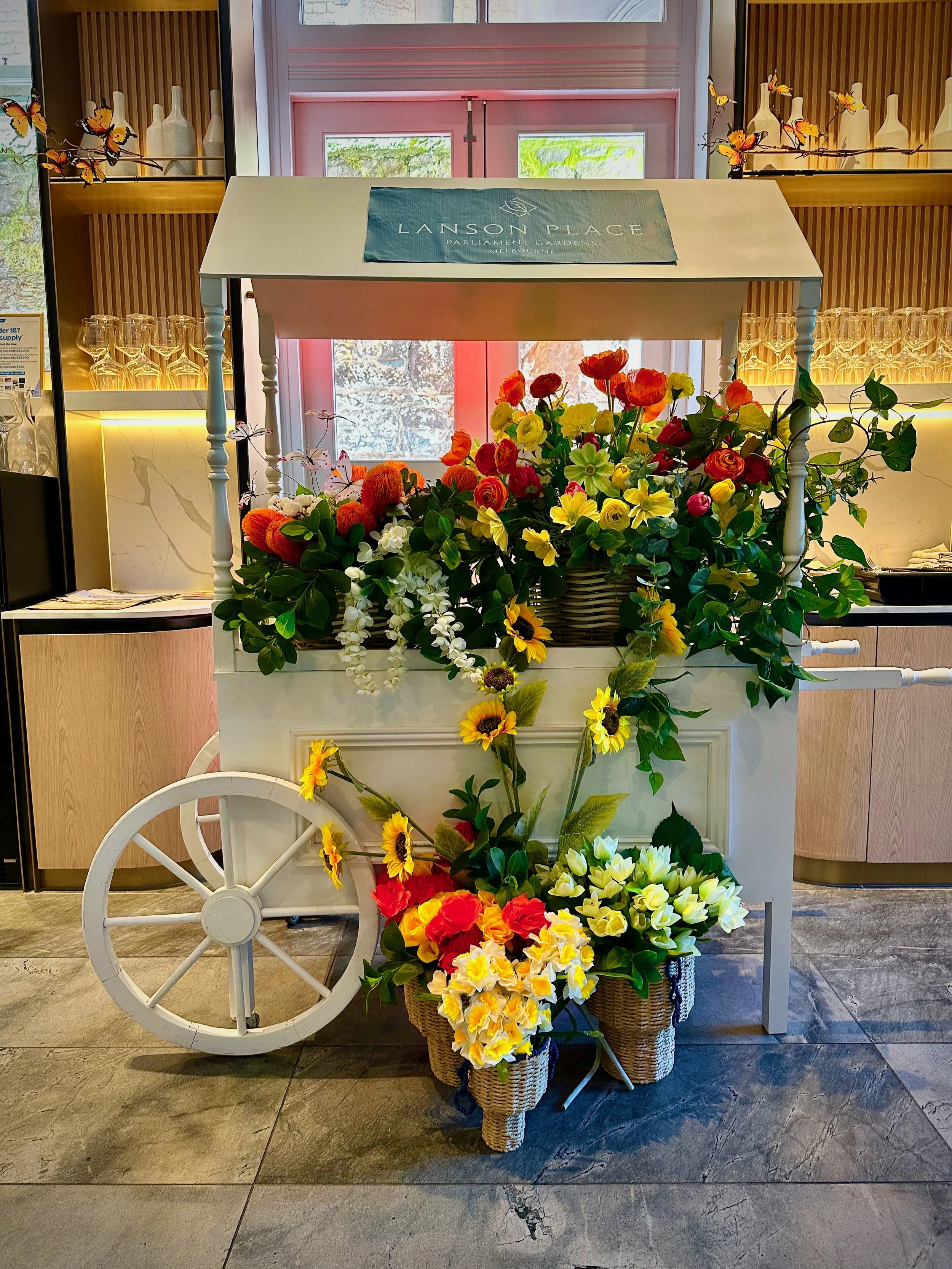 A white cart with a large bouquet of colorful artificial flowers, including sunflowers, roses, and other blooms, in front of a wooden-paneled wall with a sign that reads 'Lanson Place Parliament Gardens Melbourne' and decorative butterflies. Wicker baskets filled with additional flowers are at the base of the cart.