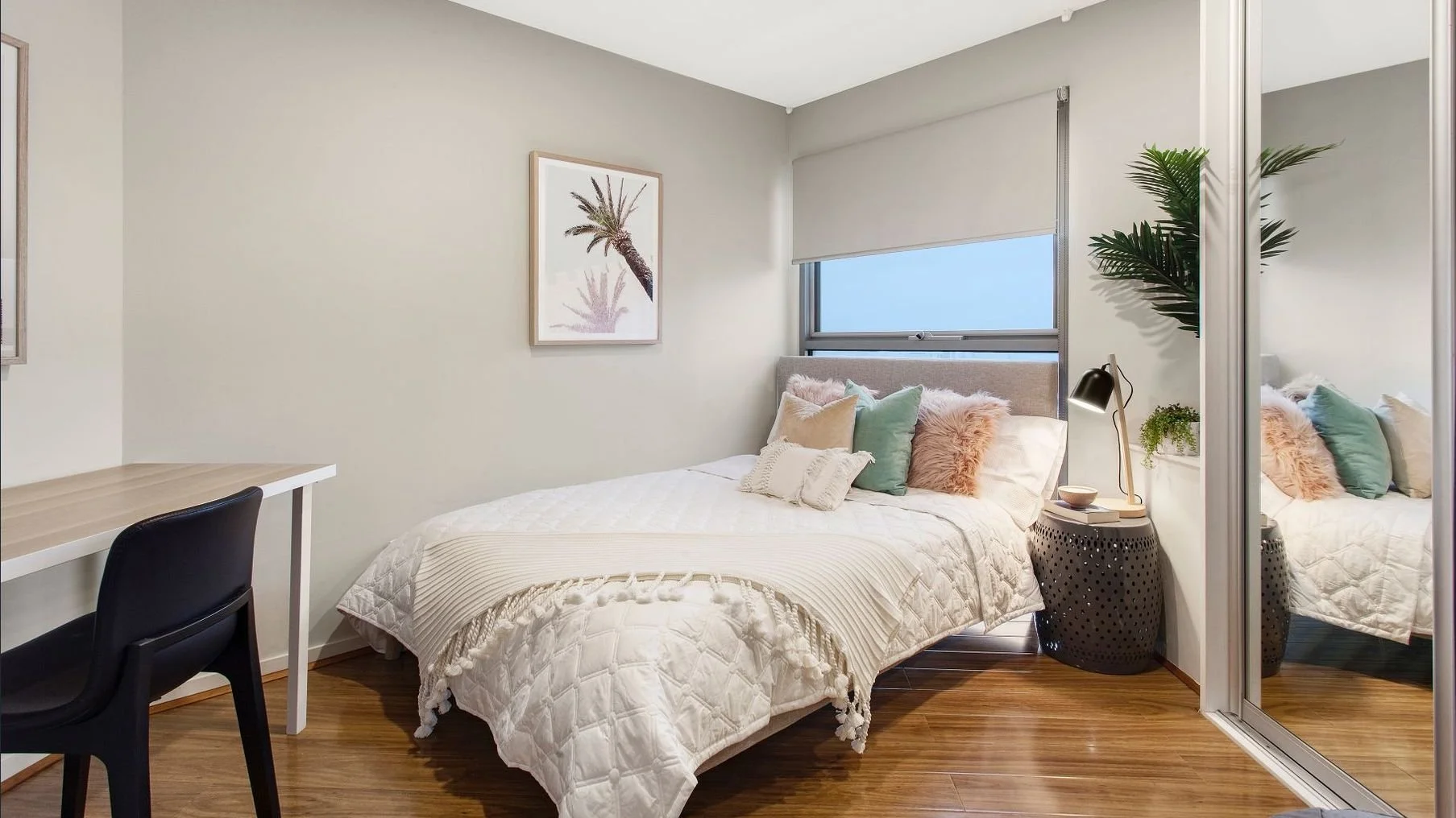 A bedroom with a bed decorated with multiple pillows, a nightstand with a lamp, a window with a shade, a wall art of a palm tree, and a mirrored closet door reflecting the bed and pillows.