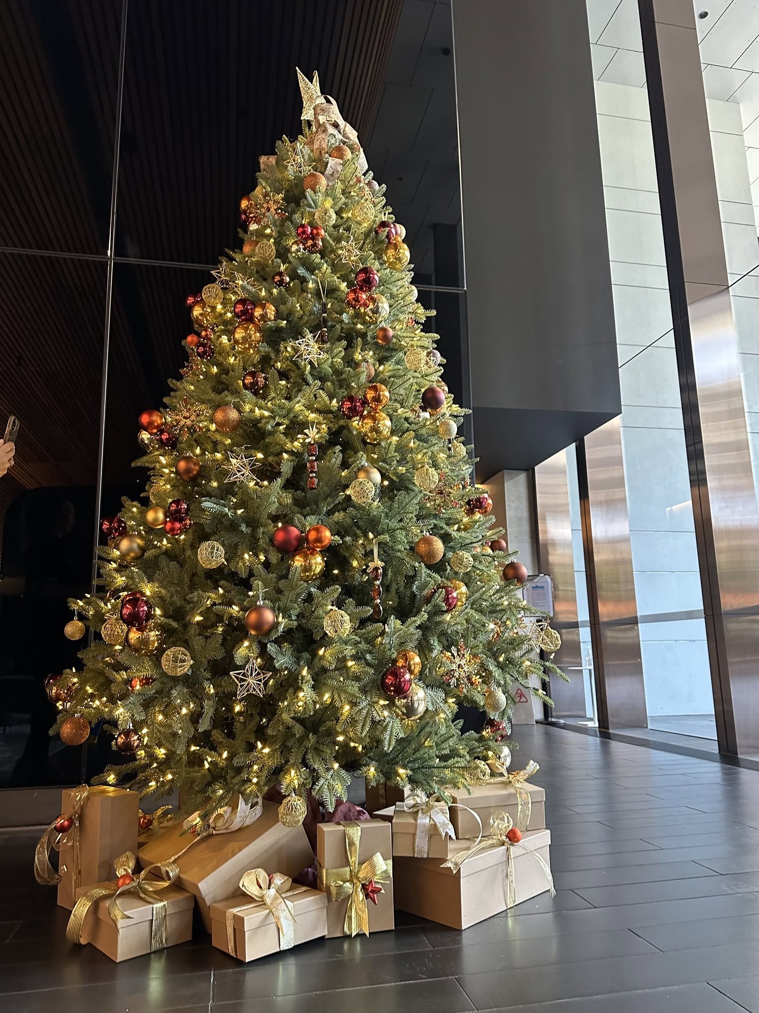 A decorated Christmas tree with wrapped gifts at the base, situated in a modern indoor space with large glass windows.