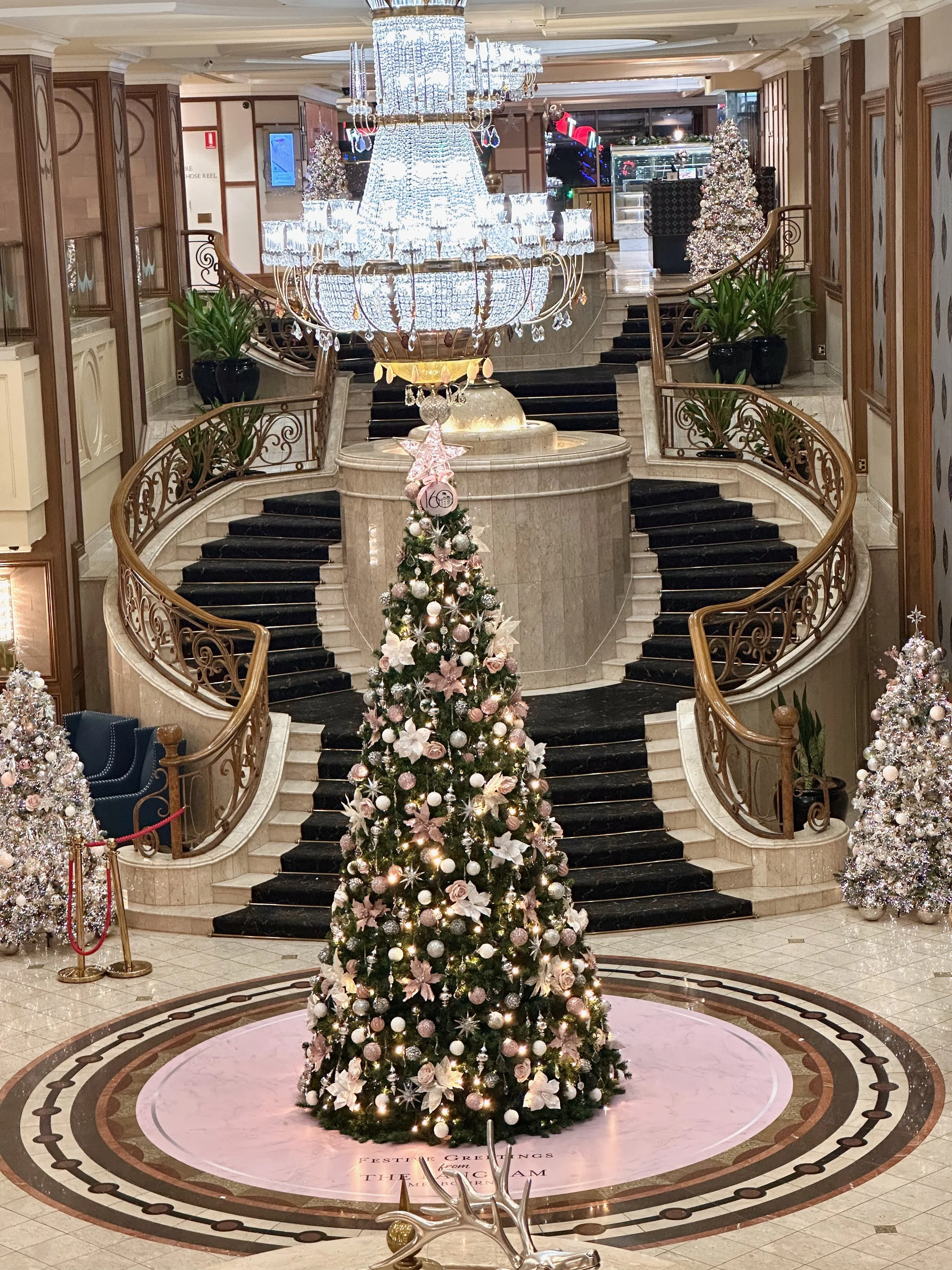 Christmas tree decorated with pink and white ornaments and flowers, surrounded by additional Christmas trees, located in an ornate hotel lobby with a grand staircase, a large chandelier, and holiday decorations.