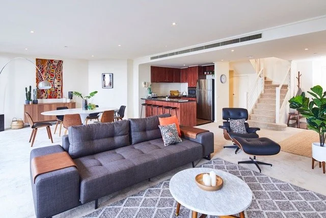 Living room and open kitchen area with modern furniture, a gray sofa, black lounge chair, round coffee table, dining table with chairs, and indoor plants.