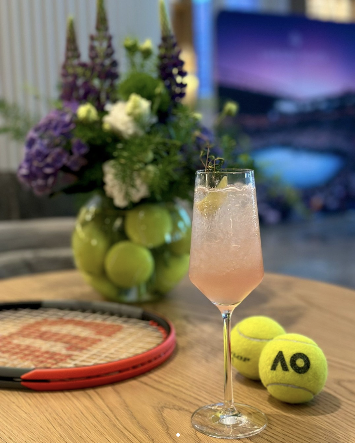 A pink cocktail in a champagne flute garnished with a lemon slice and herbs, placed on a wooden table. Behind it, there are two yellow tennis balls, a tennis racket, a bouquet of flowers in a green vase, and a blurred television screen in the backgro