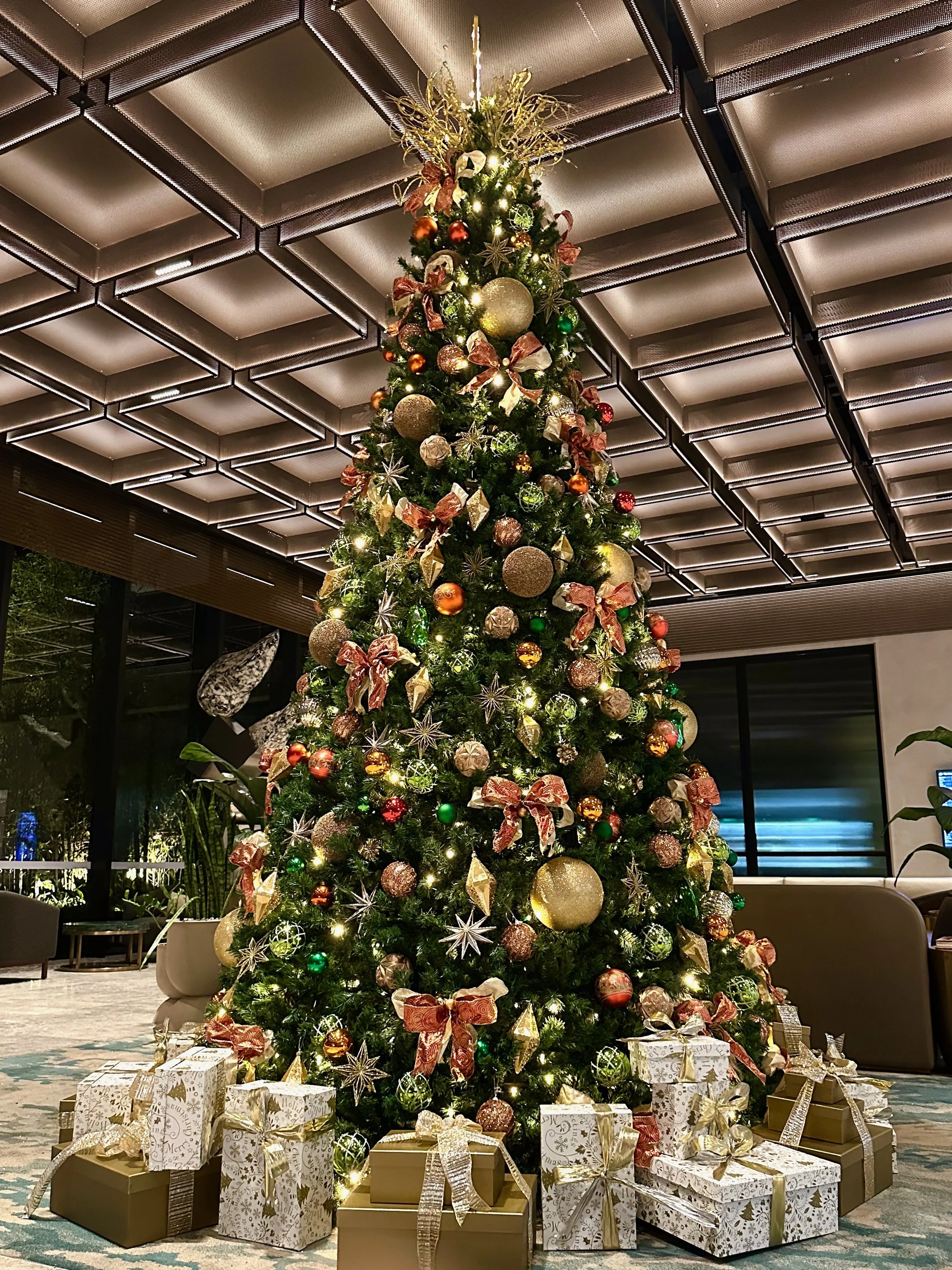 A decorated Christmas tree in the Novotel Melbourne Airport with gold, green, and red ornaments, ribbons, and lights, surrounded by wrapped presents at the base in a modern interior space.