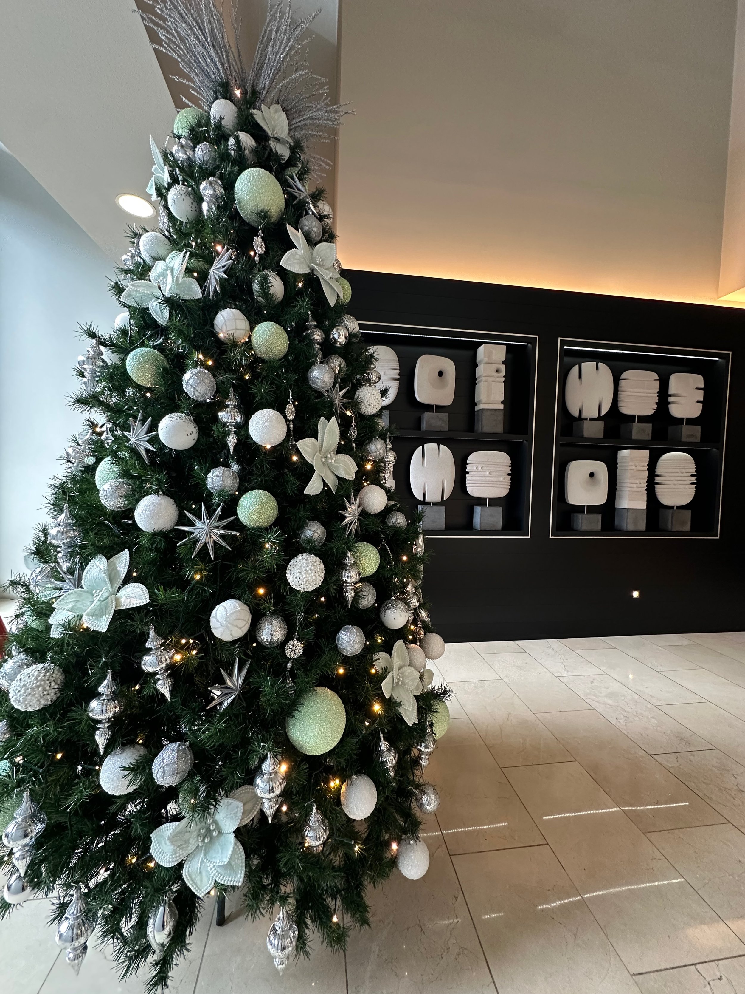 Decorated Christmas tree with white and silver ornaments, large butterfly and star decorations, and string lights in a hotel lobby area.