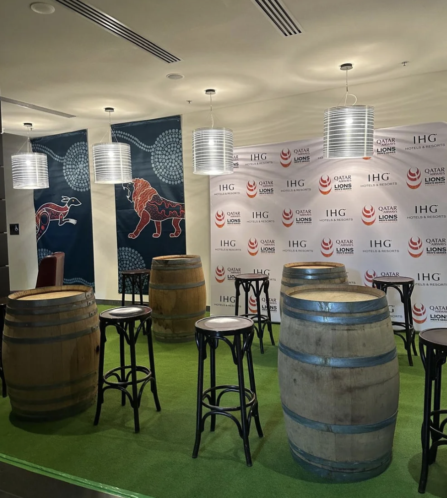 Indoor setting with a green carpet, wooden and concrete barrel tables, black barstools with white seats, hanging silver light fixtures, blue banners with camels and other designs, and a backdrop with the logos of Qatar Airways, IHGH Hotels & Resorts,