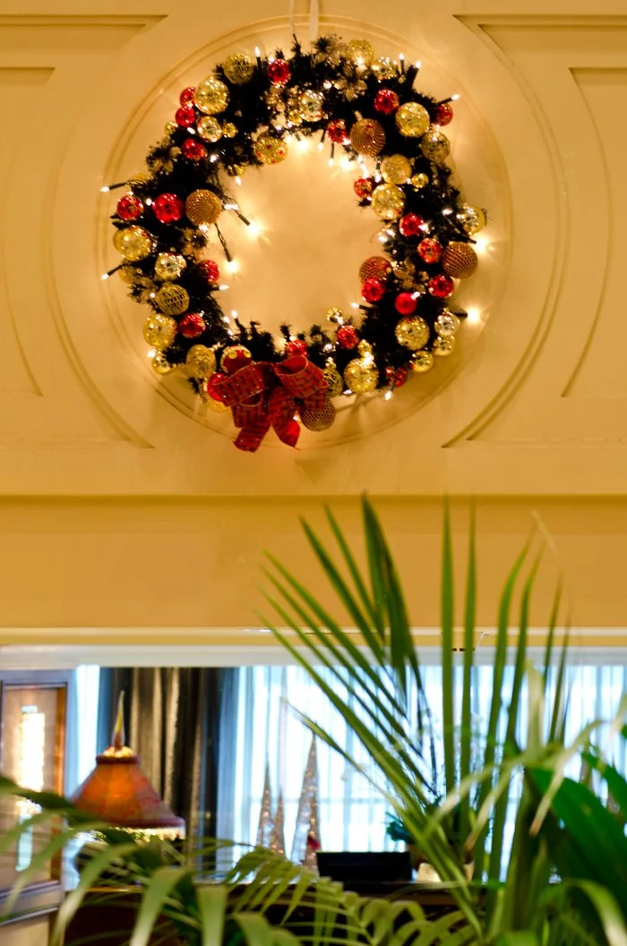 Christmas wreath with red, gold, and brown ornaments, decorated with a red bow, hanging on a wall with warm lighting.