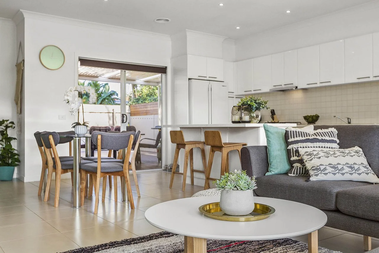 Modern living room with a gray sofa, decorative pillows, a white round coffee table with a potted plant and tray, a dining table with chairs, and a view of the outdoor patio through sliding glass doors.