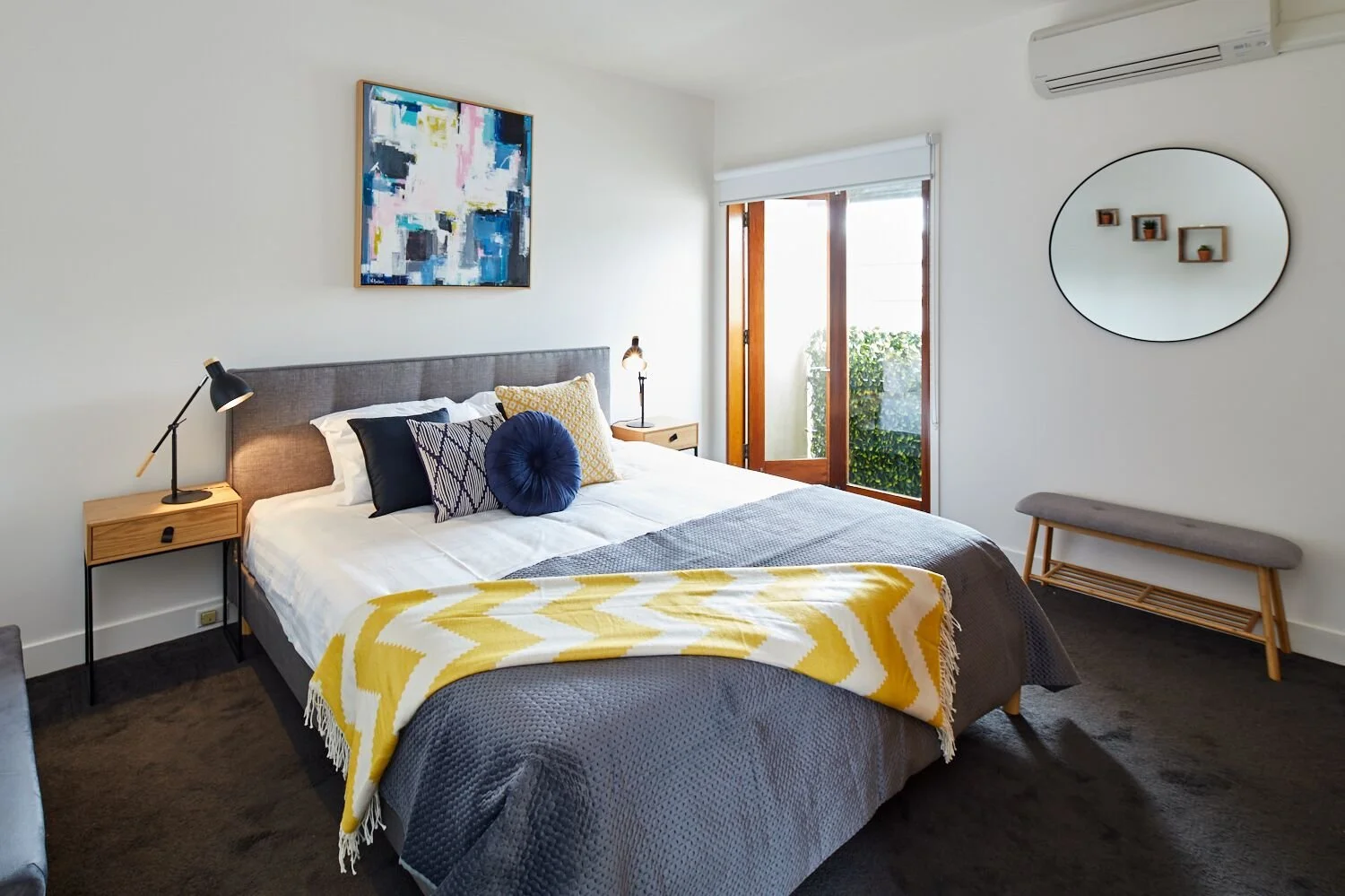 A cozy bedroom with a large bed, decorative pillows, and a yellow and white throw blanket. There are two nightstands with lamps, a colorful abstract painting above the bed, and a sliding glass door leading outside. A bench is against the wall and a r