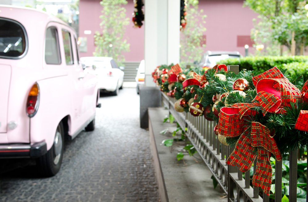 A row of Christmas wreaths decorated with red ribbons and ornaments along a metal railing  at the Langham Hotel, with parked vintage cars nearby and trees in the background.