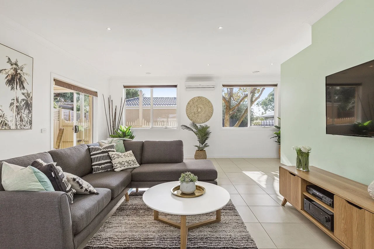 Living room with gray sectional sofa, white round coffee table, wooden TV stand, wall-mounted TV, several potted plants, abstract wall art, and large windows with blinds.