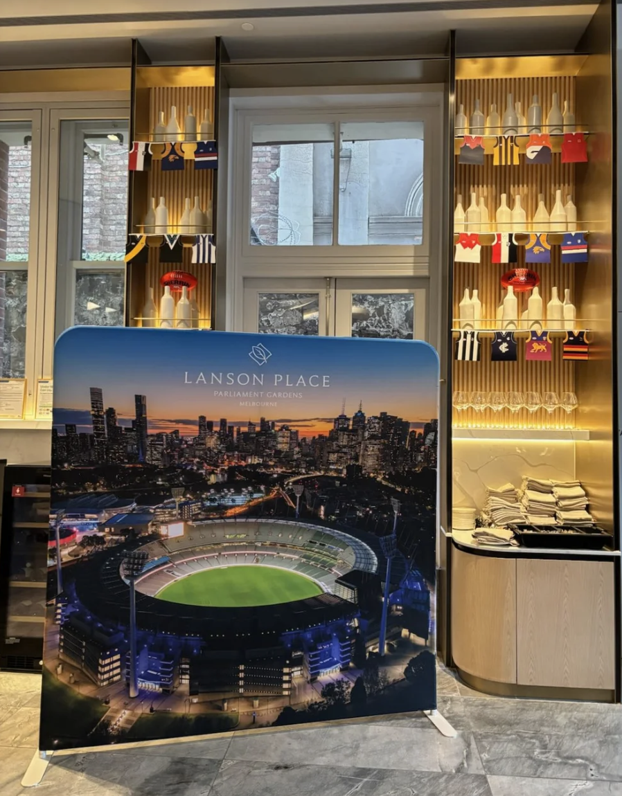 Indoor display featuring a large poster of Lanson Place Parliament Gardens in Melbourne, with a nighttime city skyline and stadium. Behind the poster, shelves show bottles and colorful flags, with stacks of paper or napkins on a counter to the right.