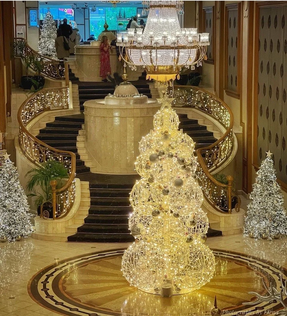 Langham Hotel lobby decorated for Christmas with a large illuminated Christmas tree made of interconnected light orbs adorned with ornaments, flanked by smaller snow-covered Christmas trees, and a grand chandelier hanging above.