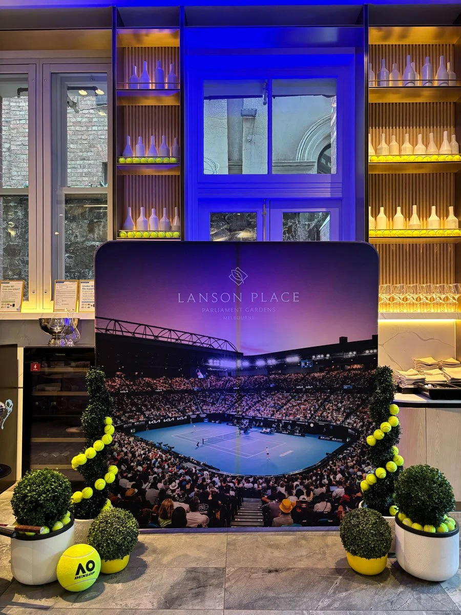Indoor area with decorative tennis-themed display promoting Lanson Place at Parliament Gardens, Melbourne, featuring a large poster of a tennis stadium, tennis balls, and potted plants.
