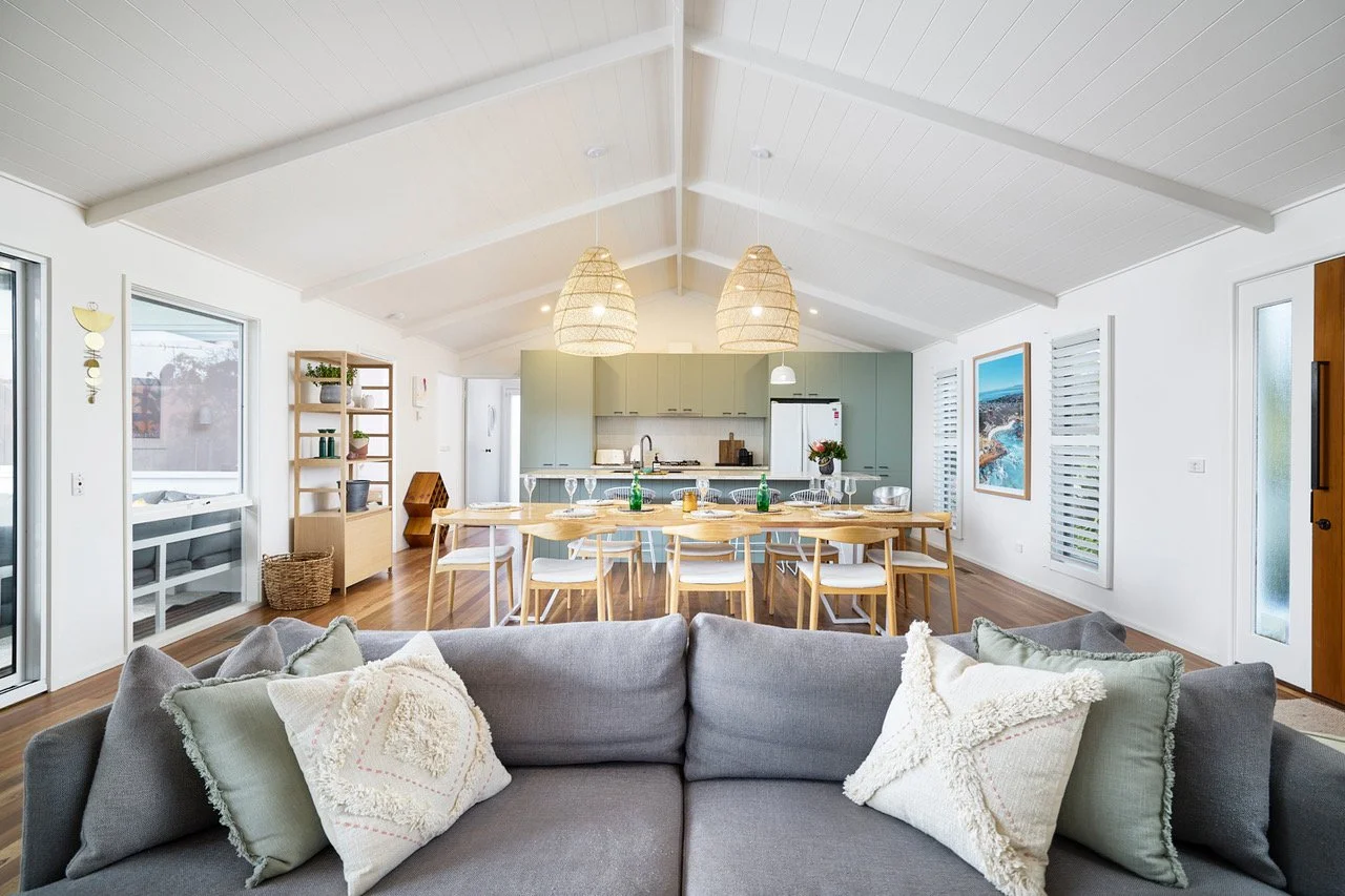 Open-concept living and dining area in a modern home with a gray sofa in the foreground, a wooden dining table, and a kitchen with light green cabinets in the background.