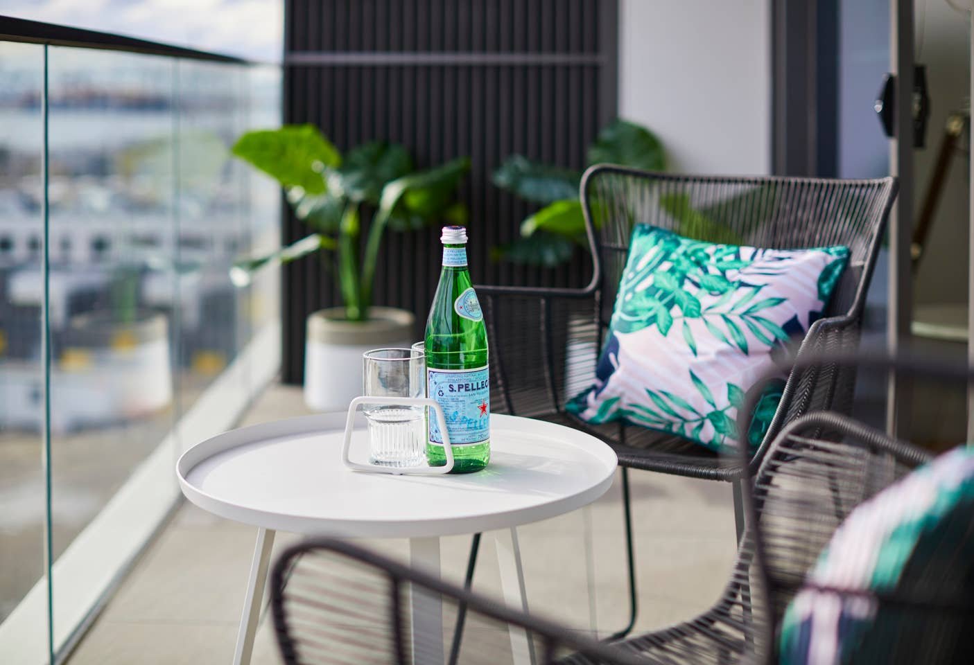 A balcony scene with a small white round table holding a green Pellegrino water bottle, an empty glass, and a glass mug. There are black wire chairs with tropical-patterned cushions, and potted plants in the background.