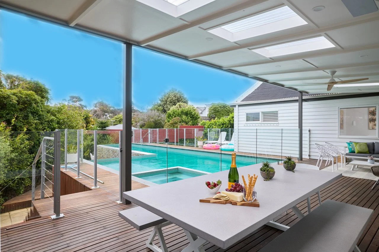 Outdoor patio area with a pool, glass fence, and seating. A table with snacks, wine, and grapes is in the foreground, with a hammock and trees in the backyard.