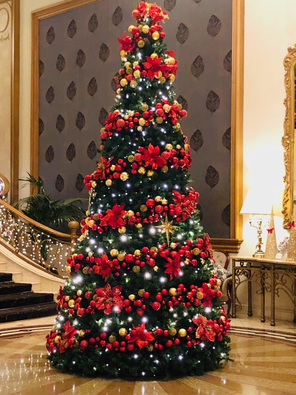 Decorated Christmas tree at the Langham Hotel with red and gold ornaments, poinsettia flowers, and white lights in a grand room with a staircase and elegant furnishings.