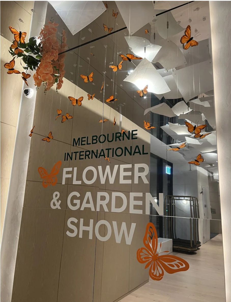 Glass wall with text promoting the Melbourne International Flower & Garden Show, decorated with orange butterflies, pink flowers, and abstract white ceiling fixtures.