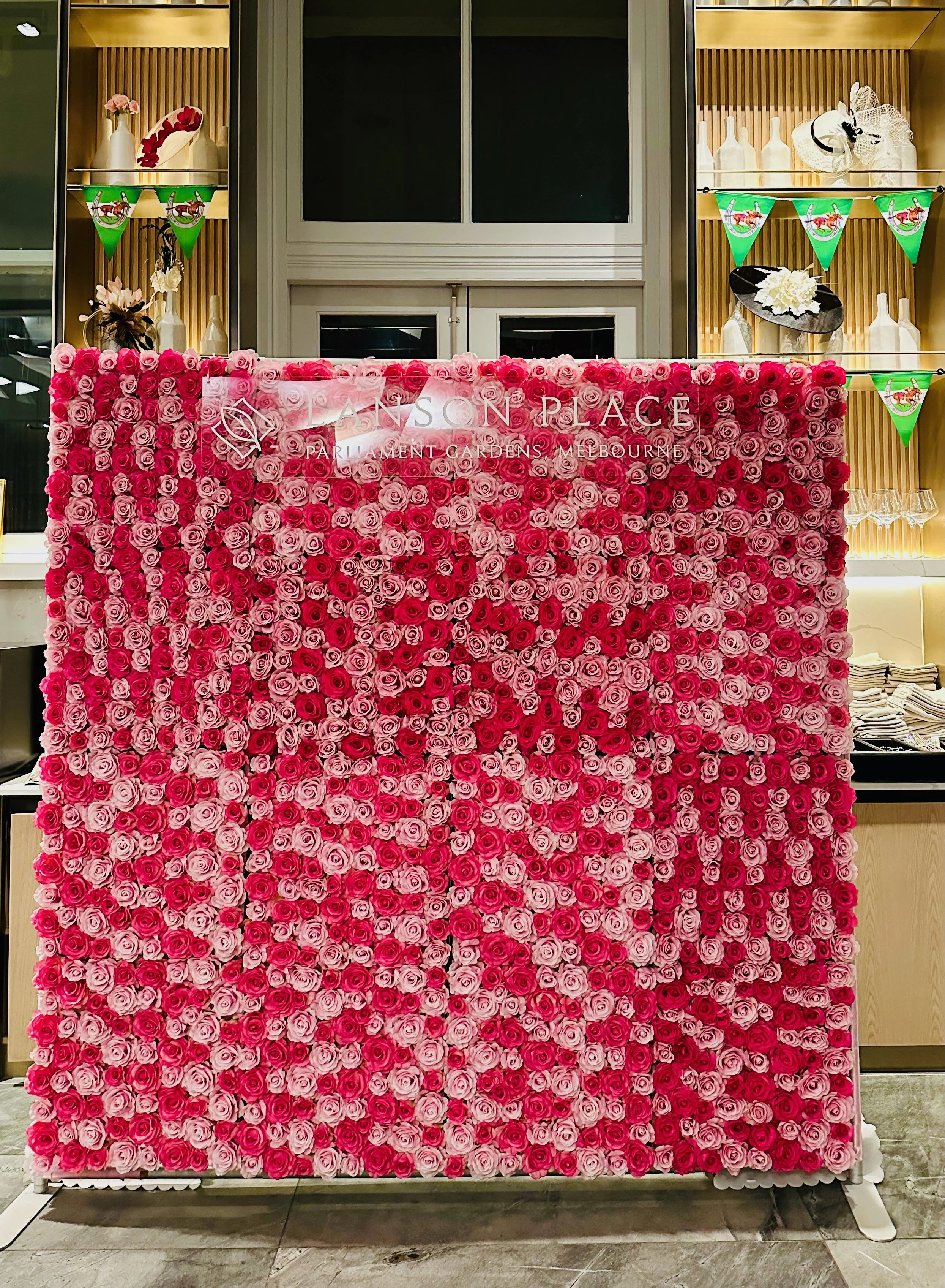 A floral display of pink and red roses in front of a wall with a sign that reads "Lanson Place Parliament Gardens Melbourne".