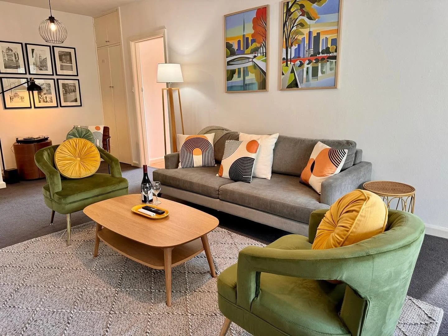A living room with a gray sofa, two green armchairs with yellow cushions, a wooden coffee table with a bottle of wine and two glasses, and decorative pillows with orange, black, and white patterns. There is a side table with a woven top, a tall floor
