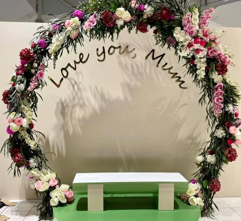 A floral arch with pink, white, and purple flowers and the words 'Love you, Mom' written in script. There is a small green and white rectangular bench in front of the arch.