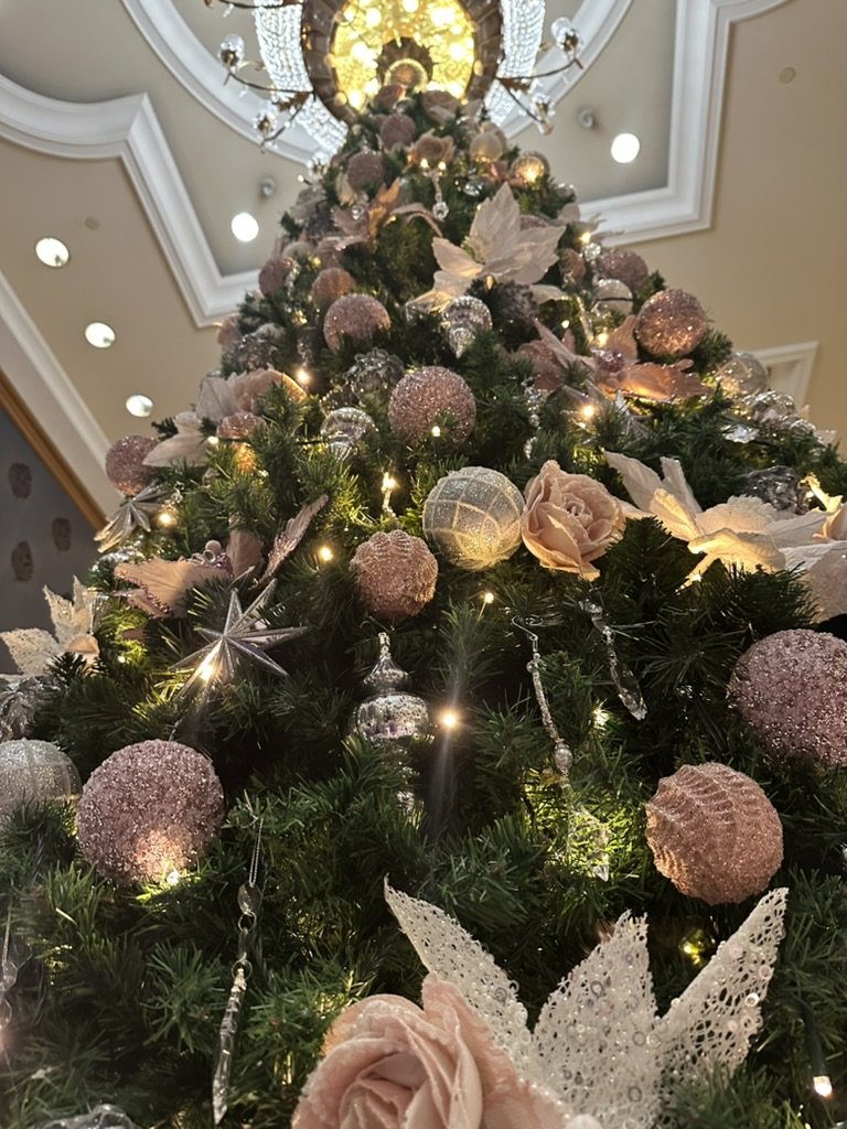 Close-up of a decorated Christmas tree with pink and silver ornaments, poinsettias, and lights, viewed from below towards a ceiling with a chandelier.