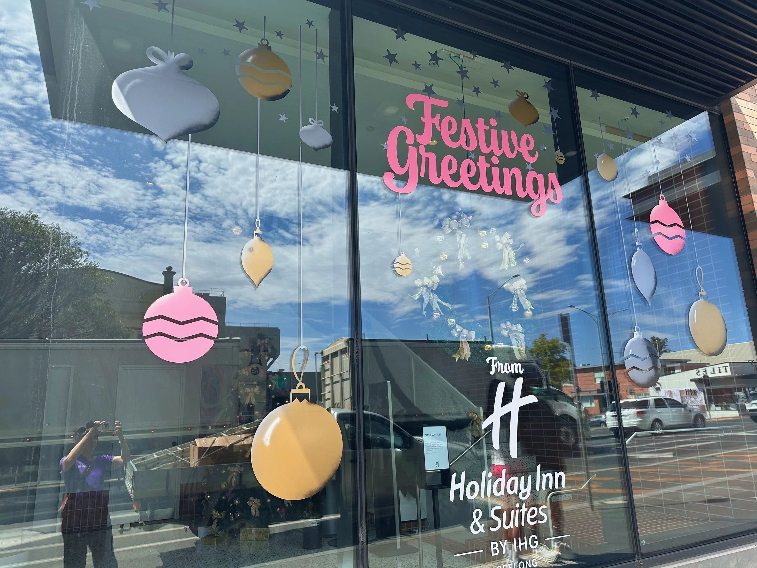 Store window decorated with Christmas ornaments and stars, with pink sign reading "Festive Greetings". Reflections of cars and a person taking the photo are visible in the glass.