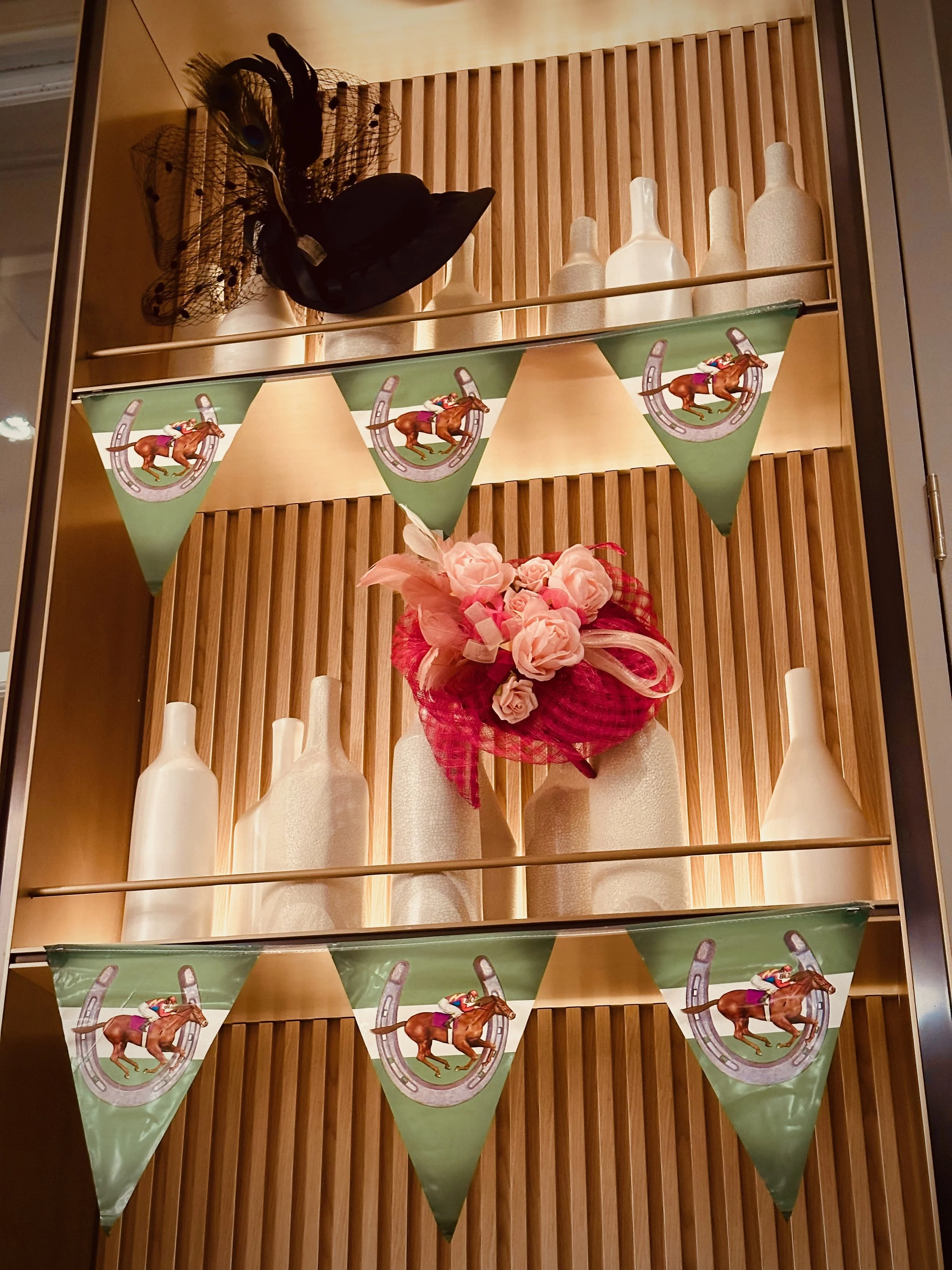 Shelf with decorative items such as a black hat adorned with feathers, ceramic vases, and a small bouquet of pink flowers. Green flags with carousel horse imagery hang in front.