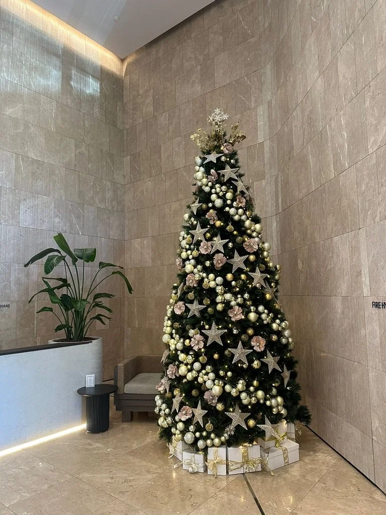 Decorated Christmas tree with white and gold ornaments, silver stars, and pink flowers, surrounded by wrapped gifts, in a modern indoor space with beige tiled walls and a plant to the side.