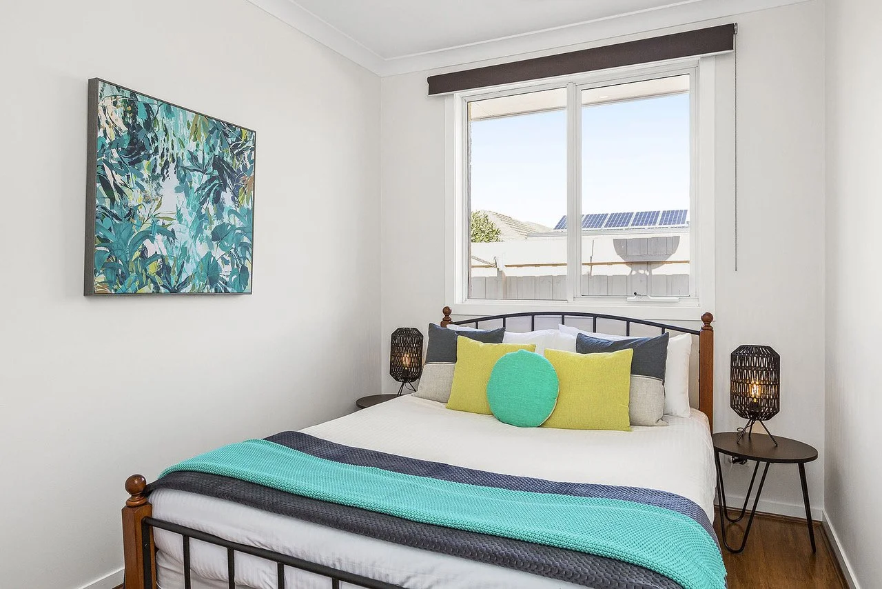 A bedroom with a white wall and a painting of green and blue leaves. There is a window with a brown valance, and outside the window are rooftops with solar panels. The bed has a white bedspread, a variety of pillows in green, gray, and white, and a t