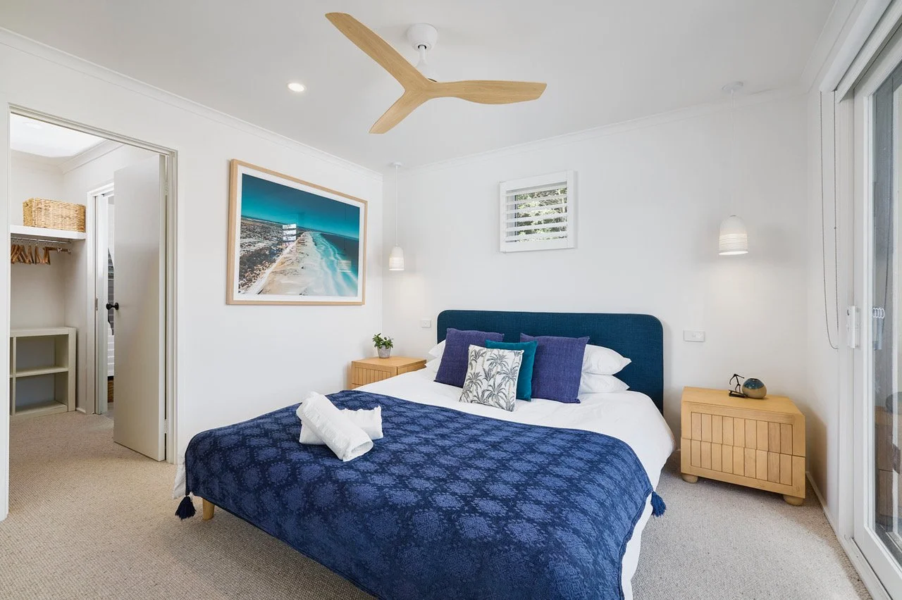 Bright bedroom with a blue upholstered bed, white bedding, and purple pillows. Light wood nightstands with a plant and a clock, ceiling fan, and sliding glass door.