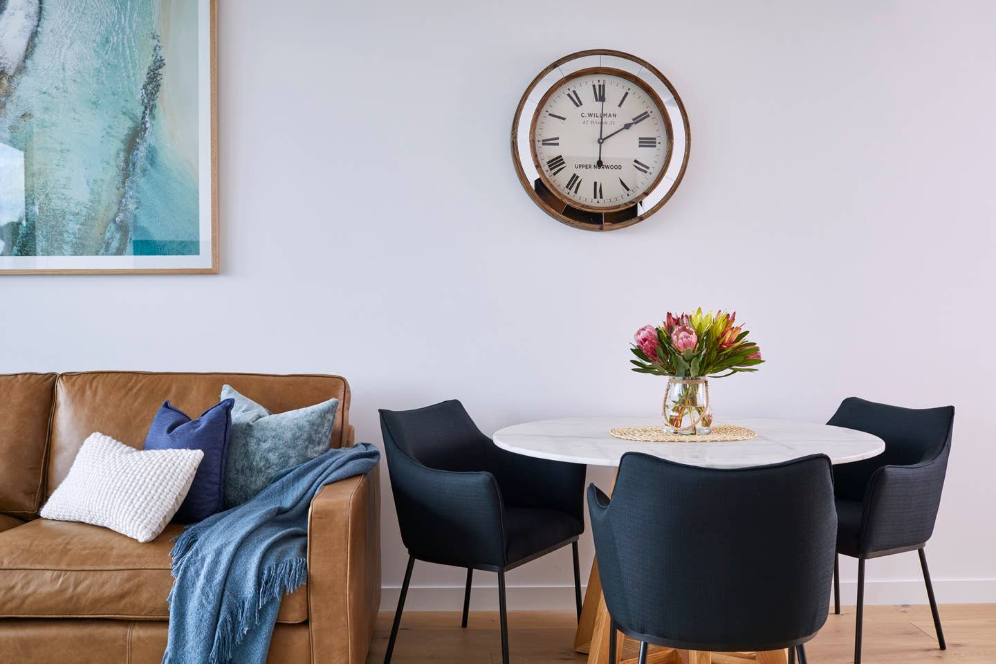 Living room with a tan leather sofa, several pillows in blue and white, a blue throw blanket, a round white table with a vase of pink flowers, and four black chairs. A modern wall clock with Roman numerals hangs above the table on a white wall.