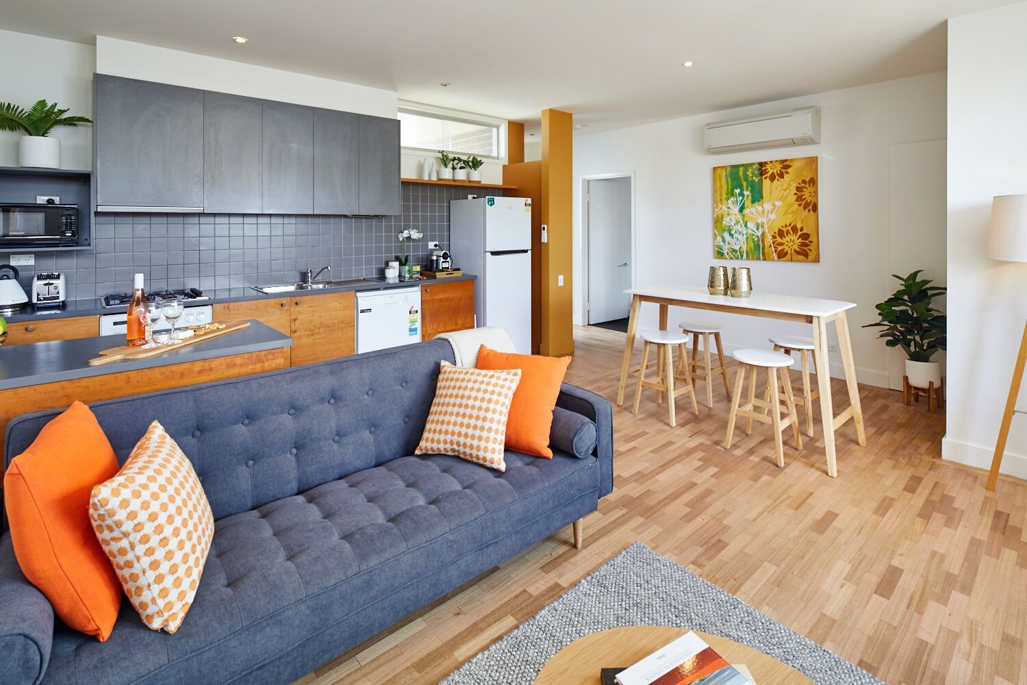 Open-concept living area with a blue sofa, orange pillows, a wooden kitchen counter, gray tile backsplash, white appliances, a dining table with stools, artwork, and houseplants.
