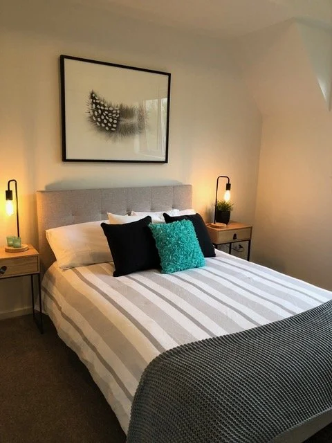 A neatly made bed with a striped beige and white bedspread, three black pillows and one turquoise pillow, flanked by two wooden nightstands with black frames and small lamps, and a large black and white feather artwork hanging above the bed.