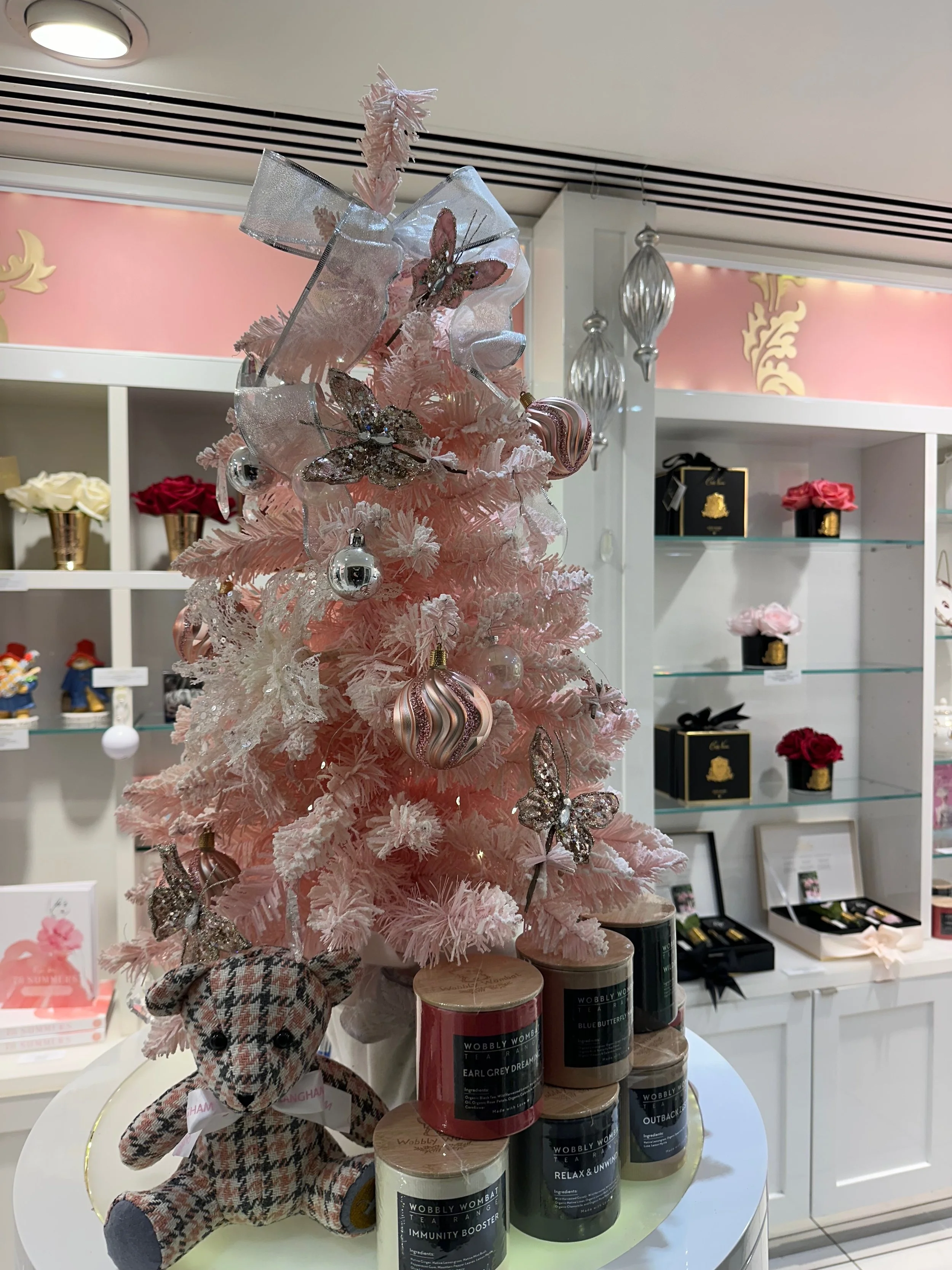 A pink Christmas tree decorated with silver butterflies, ornaments, and ribbons, in a gift shop. A plaid stuffed animal with a ribbon and tag is placed in front of the tree. There are shelves with flowers, boxes, and candles in the background.