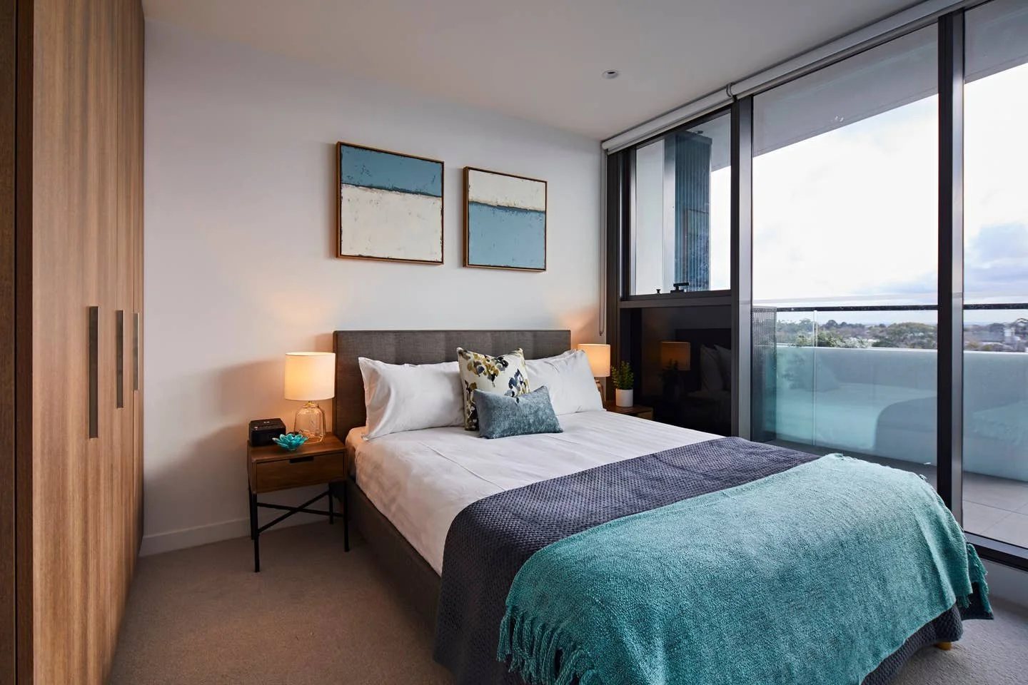 Modern bedroom with a king-sized bed, nightstands with lamps, wooden closet, and large floor-to-ceiling windowed balcony with city view.