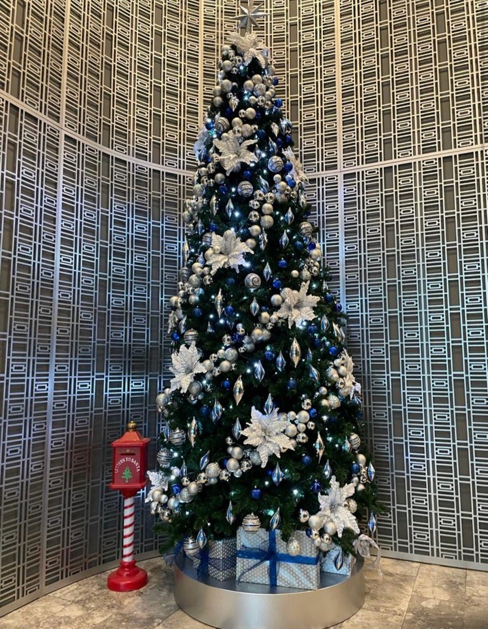 A large Christmas tree decorated with silver, blue, and white ornaments and poinsettias, with wrapped gifts at the base, in a modern interior space with patterned metal wall panels and a small red Christmas-themed mailbox nearby.