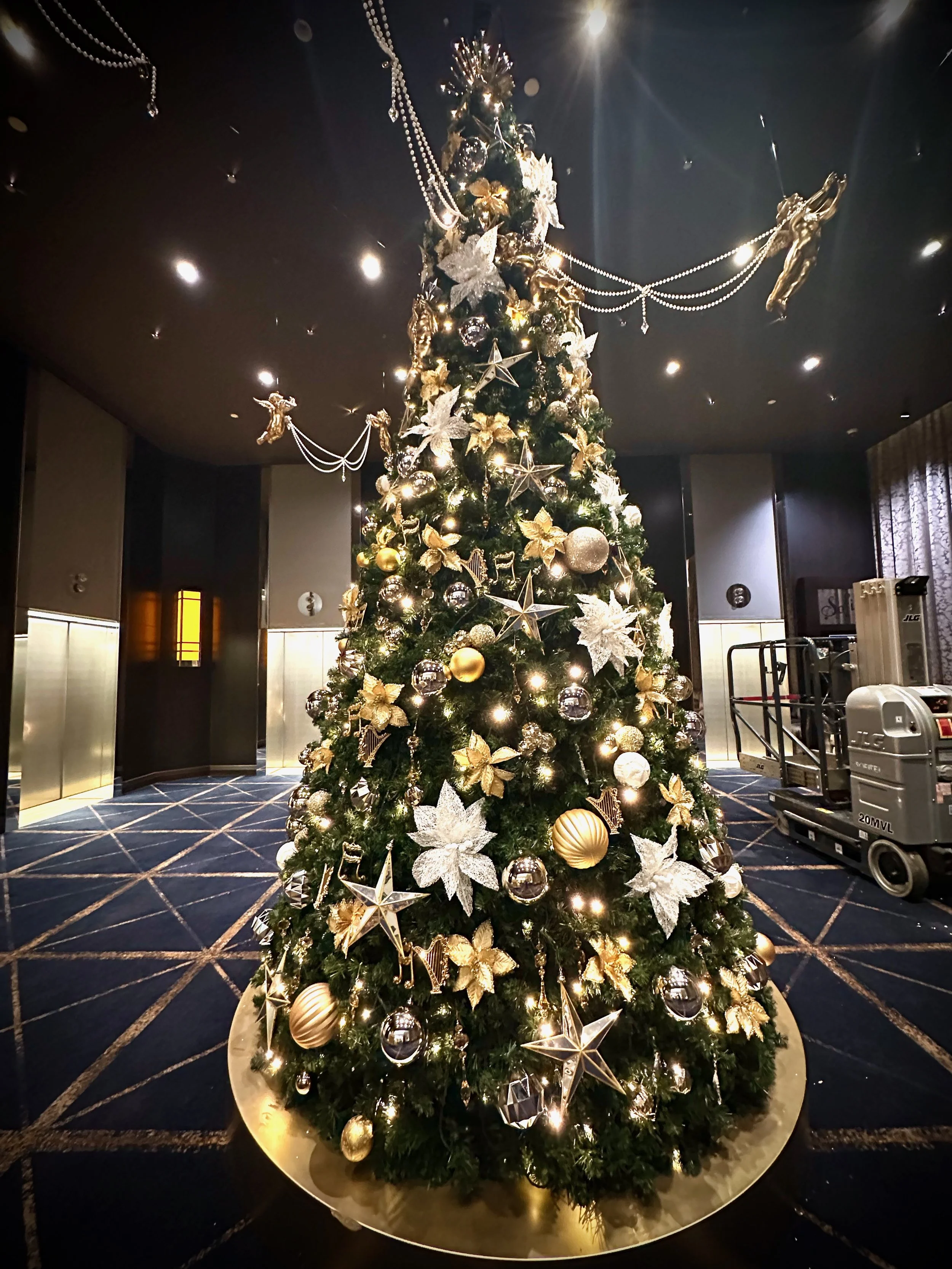 A decorated Christmas tree at Sofitel Melbourne  adorned with gold and silver ornaments, poinsettia flowers, star and angel decorations, and string lights inside a building with dark walls and a patterned carpet.