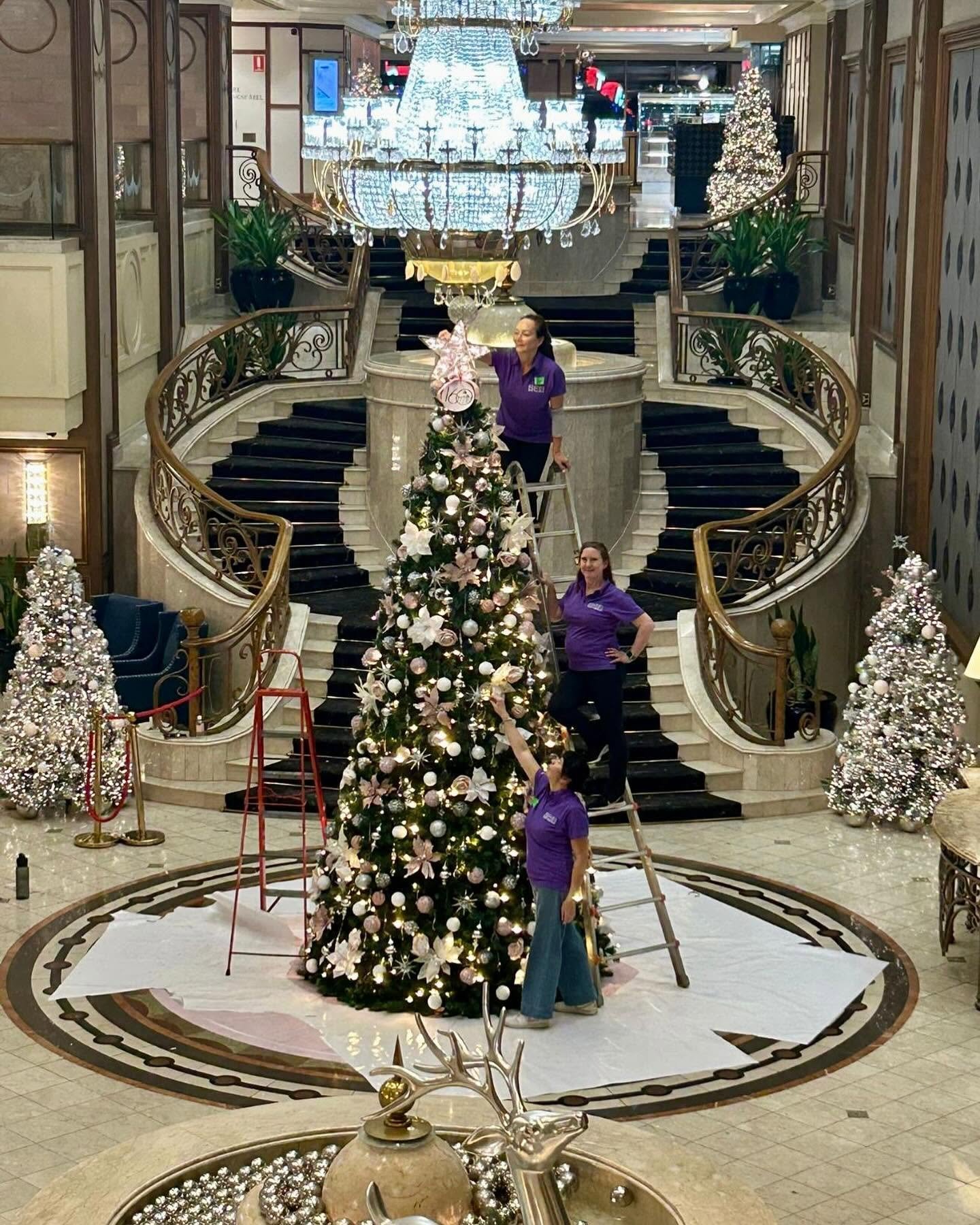 Langham Hotel lobby decorated for Christmas with large Christmas tree, smaller trees, and workers decorating. Elegant staircase and chandelier in the background.