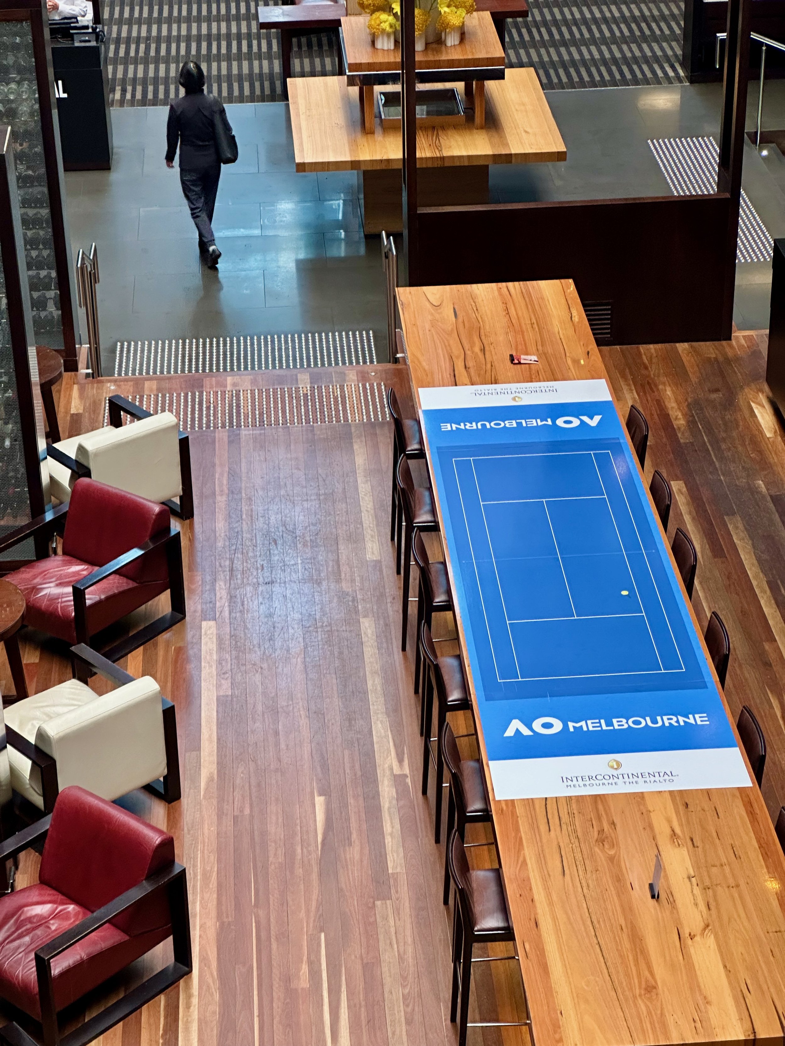 An indoor scene showing a wooden conference table with a blue tennis-themed table cover featuring the AO Melbourne logo, surrounded by chairs. In the background, a woman is walking away from the camera, carrying a tote bag, with wooden tables decorat