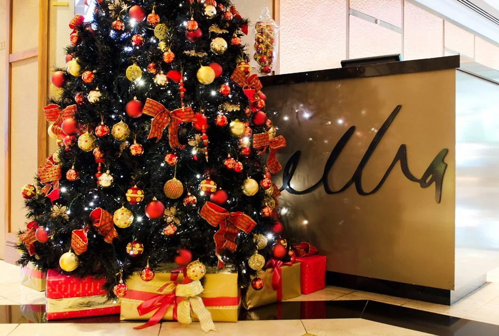 Decorated Christmas tree at Melba Restaurant with red, gold, and white ornaments, red ribbons, and gift boxes underneath, located next to a sign with the word 'Ulls' on it.