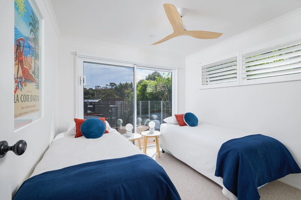 A bright bedroom with two white beds, each with a blue blanket and colorful pillows, a sliding glass door leading to a balcony, and a ceiling fan with wooden blades.